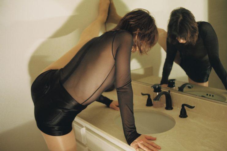 A dancer leaned over a bathroom sink with her left leg on the wall.